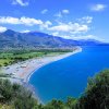 Vista aerea di una costa con spiaggia e mare blu, circondata da montagne e vegetazione.